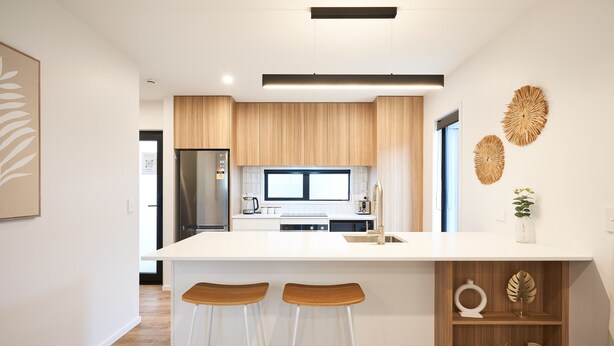 A modern kitchen with a central island, wooden cabinets, and a white countertop. There are two bar stools and decorative items on the island. - Newly built unit in St Kilda. Modern amenities (Dunedin)
