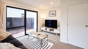 A modern living room with a sofa, a glass coffee table, a flat-screen TV, and a sliding glass door leading to an outdoor area. - Newly built unit in St Kilda. Modern amenities (Dunedin)