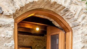 Une chambre aux murs de pierre, avec une porte en bois, un lit et une table de chevet sur laquelle se trouve une lampe.