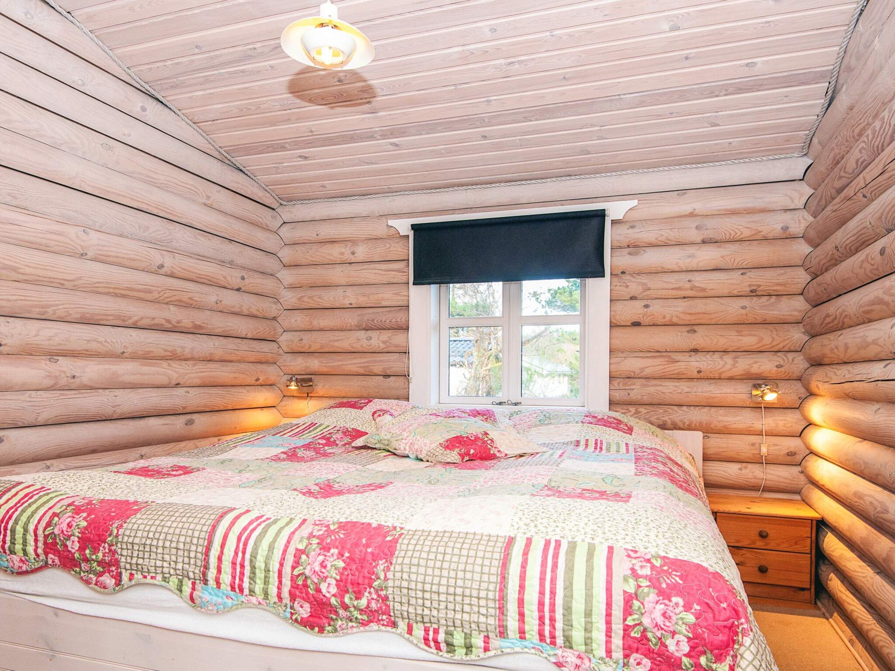 A log cabin room with a bed, a window with a black valance, and two bedside lamps.