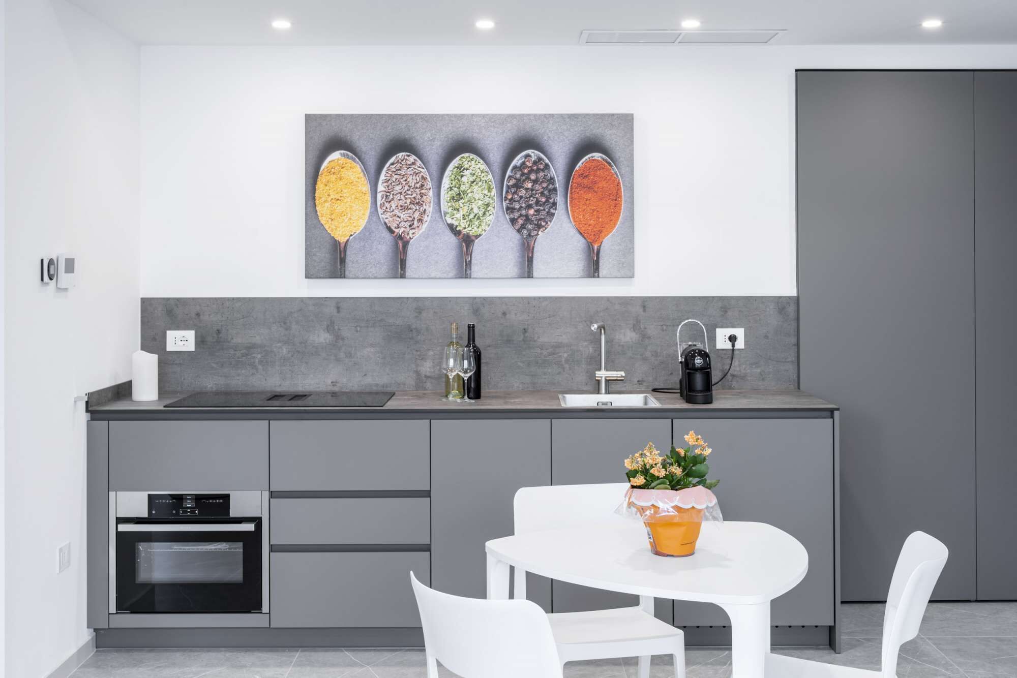 A modern kitchen with a grey countertop, a built-in oven, and a coffee machine. There is a dining table with chairs and a vase of flowers.