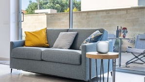 A grey sofa with mustard and patterned cushions, a small round wooden table with a white vase, and a view of a patio area with outdoor furniture and a brick wall.