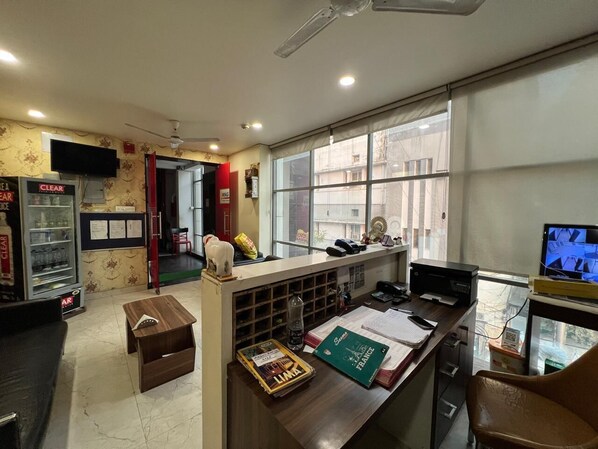 A reception area with a counter, a refrigerator, a television, and a window with a view of buildings. - Hotel The Mall Inn by MNG Rooms (Kanpur)