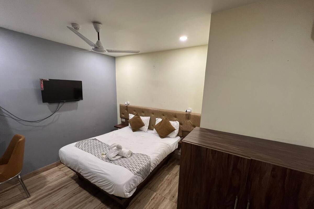 Ein ordentlich eingerichtetes Hotelzimmer mit einem Bett, einem Nachttisch aus Holz, einem an der Wand montierten Fernseher und einem Deckenventilator.