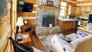 A log cabin interior with a stone fireplace, wooden furniture, a kitchen, and a mounted television.