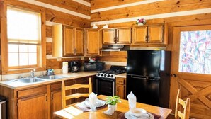 A wooden cabin-style kitchen with a dining table set for two, a stove, refrigerator, and a window with blinds.