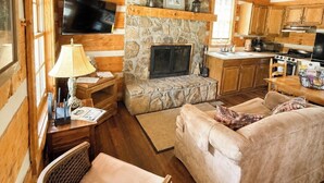 A cozy cabin interior with a stone fireplace, wooden walls, a television, a dining table, and a sofa.