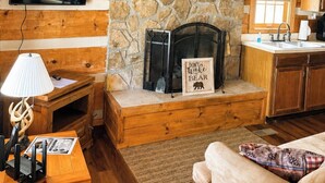 Un interior de cabaña acogedor con chimenea de piedra, paredes de madera, televisor montado y kitchenette.