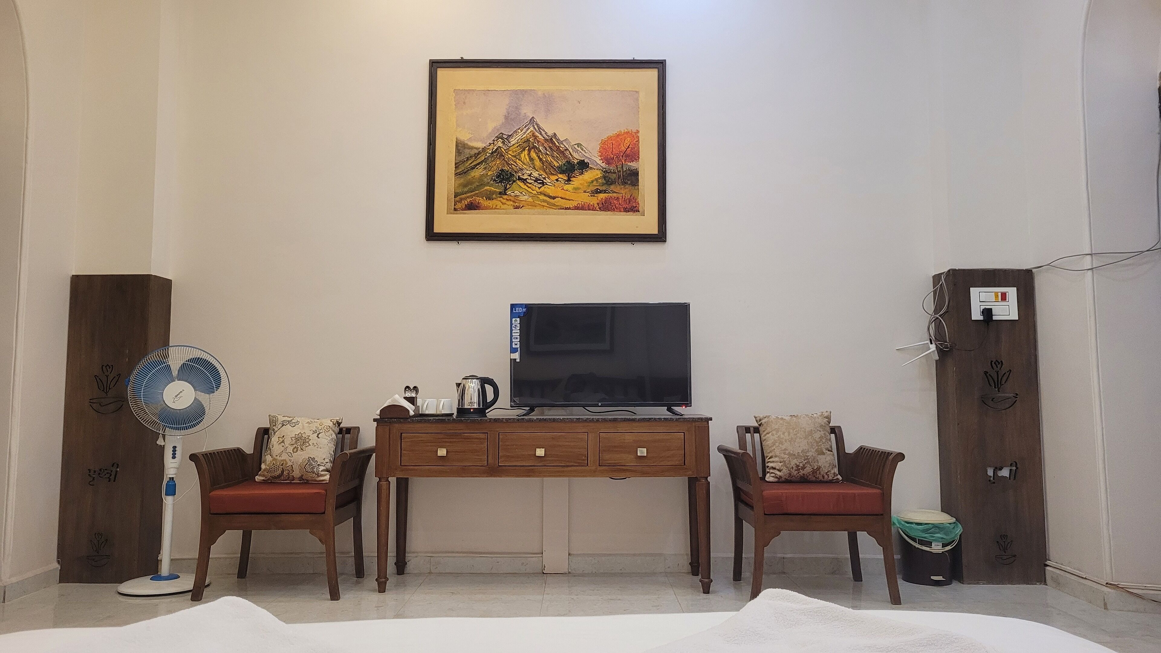 Un soggiorno con una TV a schermo piatto su un supporto in legno, due poltrone di legno con cuscini, una ventola a piantana e un quadro appeso al muro.
