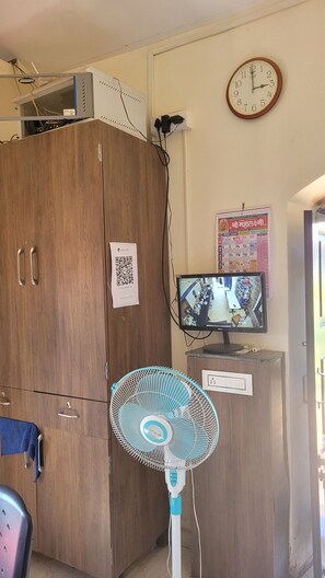 A room with a wooden wardrobe, a wall-mounted clock, a television, and a fan. - Madhuvan Retreat Resort Amba (Shahuwadi)