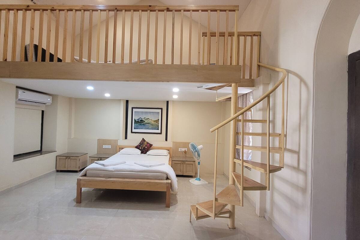 Un dormitorio con literas, un escritorio, una silla, una cama con almohadas, una mesita de noche, aire acondicionado instalado en la pared, un cuadro enmarcado y una escalera de caracol.