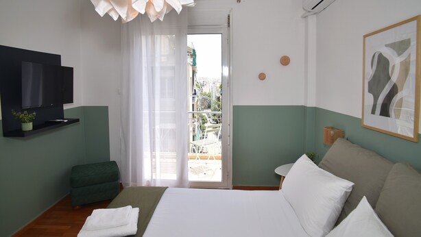 A hotel room with a bed, a TV, a green ottoman, a balcony with a view, and a painting on the wall. - Acropolis Ifigenia Sunny 2 BR, Koukaki, Steps from Landmarks (Athina)
