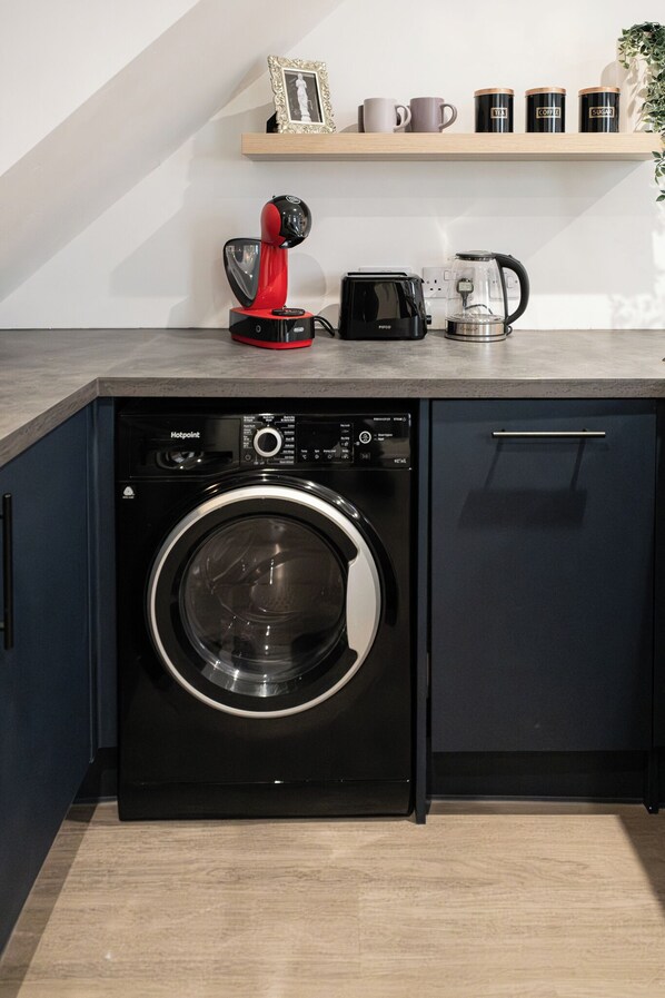 Cocina compacta con lavadora de carga frontal, cafetera roja y cafetera sobre la encimera.