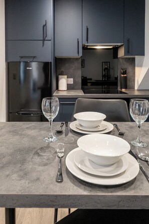 A modern kitchen with dark cabinets, a stainless steel refrigerator, and a dining table set with white plates, bowls, and wine glasses. - Gorgeous 1 BD in Central Cheltenham (Cheltenham)