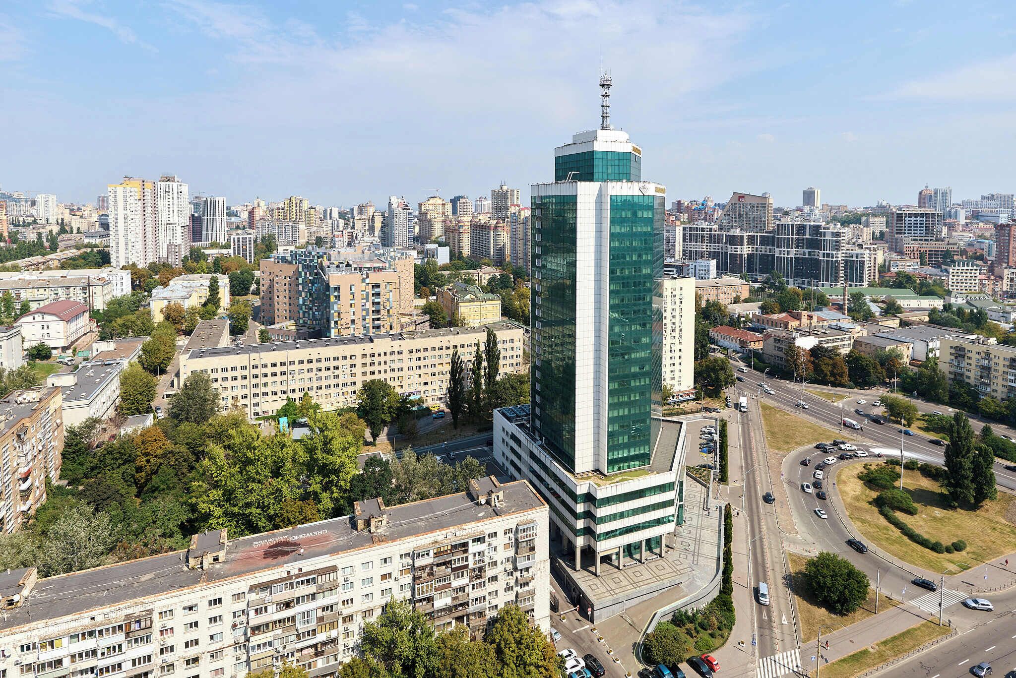 Un edificio de gran altura con fachada de vidrio moderna, rodeado de edificios residenciales más bajos y una calle concurrida con vehículos.