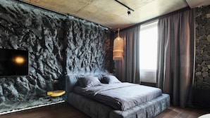 Une chambre avec un mur en pierre, un lit avec une literie grise, un luminaire suspendu et une fenêtre avec des rideaux.