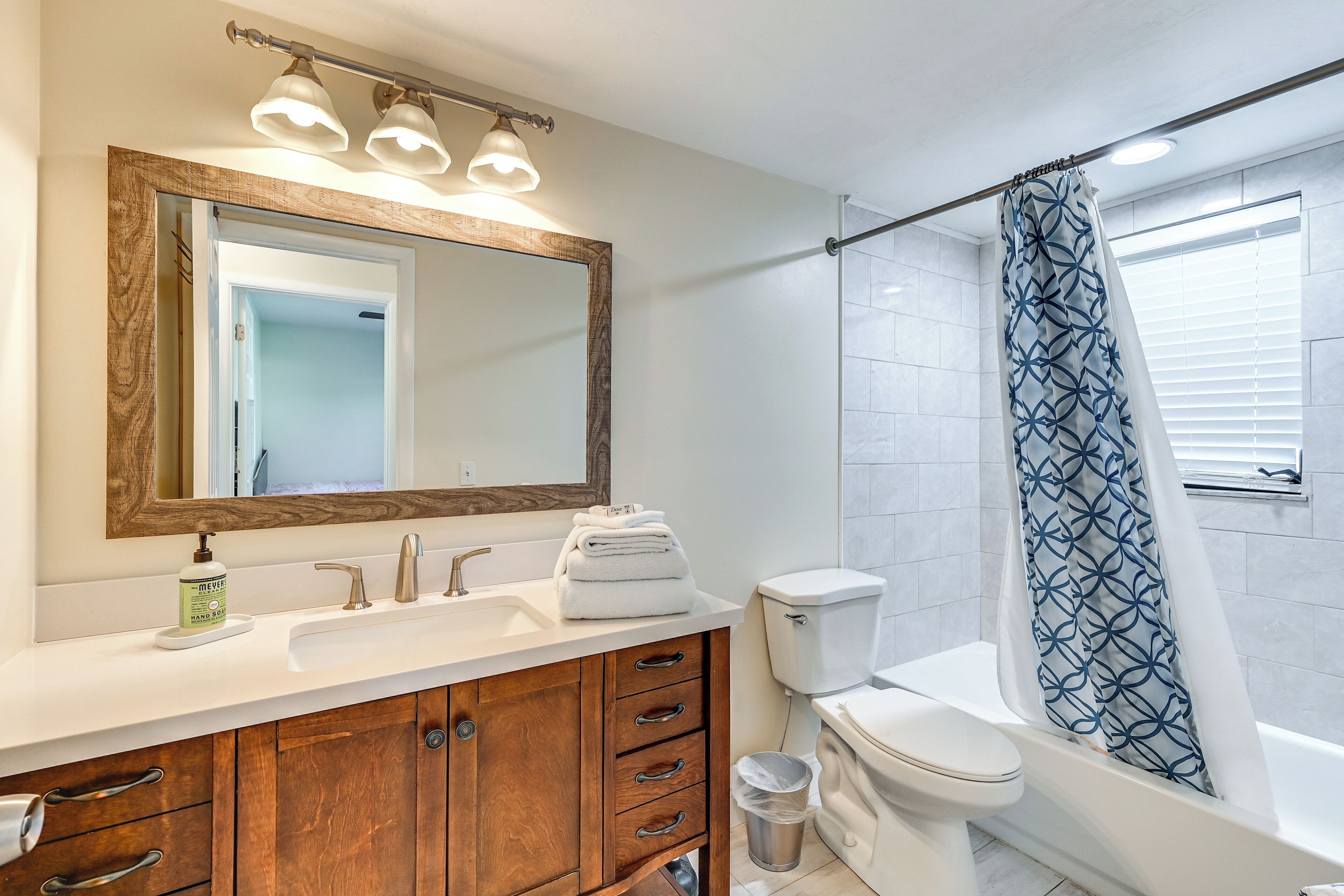 Une salle de bain avec un meuble-lavabo en bois, un grand miroir, des toilettes et une douche dotée d’un rideau à motifs.