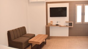 Un salon moderne avec un canapé marron, une table basse en bois, un téléviseur à écran plat fixé au mur et un petit coin cuisine doté d’un plan de travail et de placards.