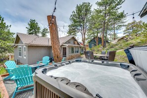 A hot tub with a stone surround, outdoor seating with Adirondack chairs, and a view of a house with a stone chimney. - Steps to Estes Lake: Mtn-view Hideaway w/ Hot Tub! (Estes Park)