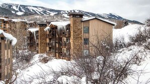 Ein mit Schnee bedecktes Gebäude mit einer Stein-Fassade und zahlreichen Fenstern, umgeben von schneebedeckten Bäumen und Bergen im Hintergrund.