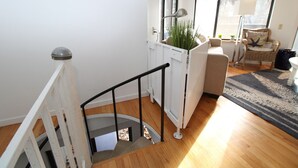 Una escalera con barandilla blanca, piso de madera, pared blanca, una ventana, una silla y una lámpara.