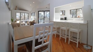 Amplia sala de estar con una gran mesa de comedor de madera, sillas blancas y una cocina con armarios y encimera blancos.