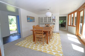 A dining area with a wooden table and chairs, a yellow patterned rug, and a view of the outdoors through a door. - Spacious Home With Private Yard Close to Beach Abutting Conservation (2397) (Brewster)