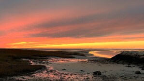 Un paysage côtier au coucher du soleil, avec une plage de sable, des zones herbeuses et un promontoire rocheux.
