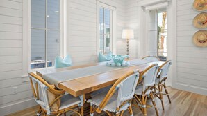 A dining area with a wooden table and wicker chairs, white paneled walls, and a view through a window.