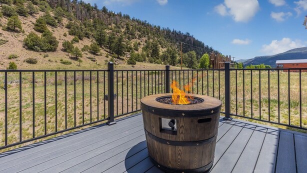 A deck with a barrel fire pit surrounded by a black metal fence, overlooking a hilly landscape with trees and a clear sky. - Newly Built Duplex | A/C | Peekaboo River Views | Cozy Interior (South Fork)
