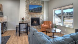 A modern living room with a stone fireplace, a large flat-screen TV mounted on the wall, a comfortable sofa, a small round table, and a window with a view of a landscape. - Newly Built Duplex | A/C | Peekaboo River Views | Cozy Interior (South Fork)