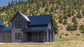 A two-story house with a dark blue roof, situated in a rural area with a rocky hillside and sparse vegetation in the background. - Newly Built Duplex | A/C | Peekaboo River Views | Cozy Interior (South Fork)