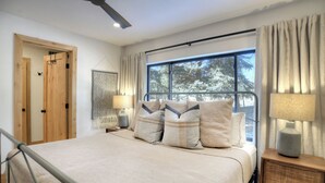 A bedroom with a large bed, two bedside tables with lamps, a window with curtains, and a wooden door.