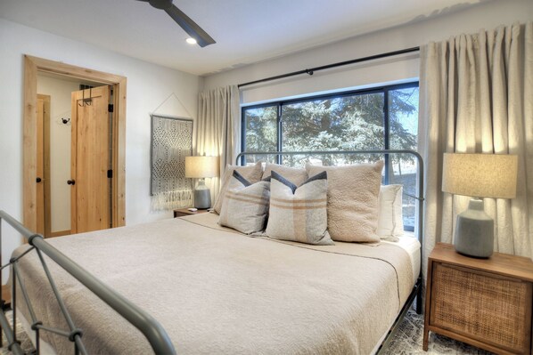 Un dormitorio con una cama grande, dos mesitas de noche con lámparas, una ventana con cortinas y una puerta de madera.