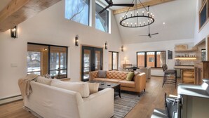 A spacious living area with a high ceiling, large windows, a central seating area with a sofa and chairs, a chandelier, and a kitchen area with wooden cabinets and a countertop.