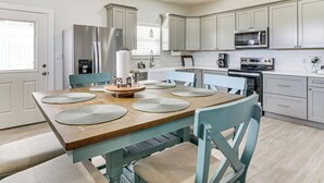 A modern kitchen with grey cabinets, a stainless steel refrigerator, a microwave, and a dining area with a wooden table and blue chairs. - Modern Port Isabel Home: Coastal Family Home! (Port Isabel)