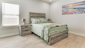 A bedroom with a wooden bed, a nightstand, a window with blinds, and a painting of a sunset on the wall. - Modern Port Isabel Home: Coastal Family Home! (Port Isabel)