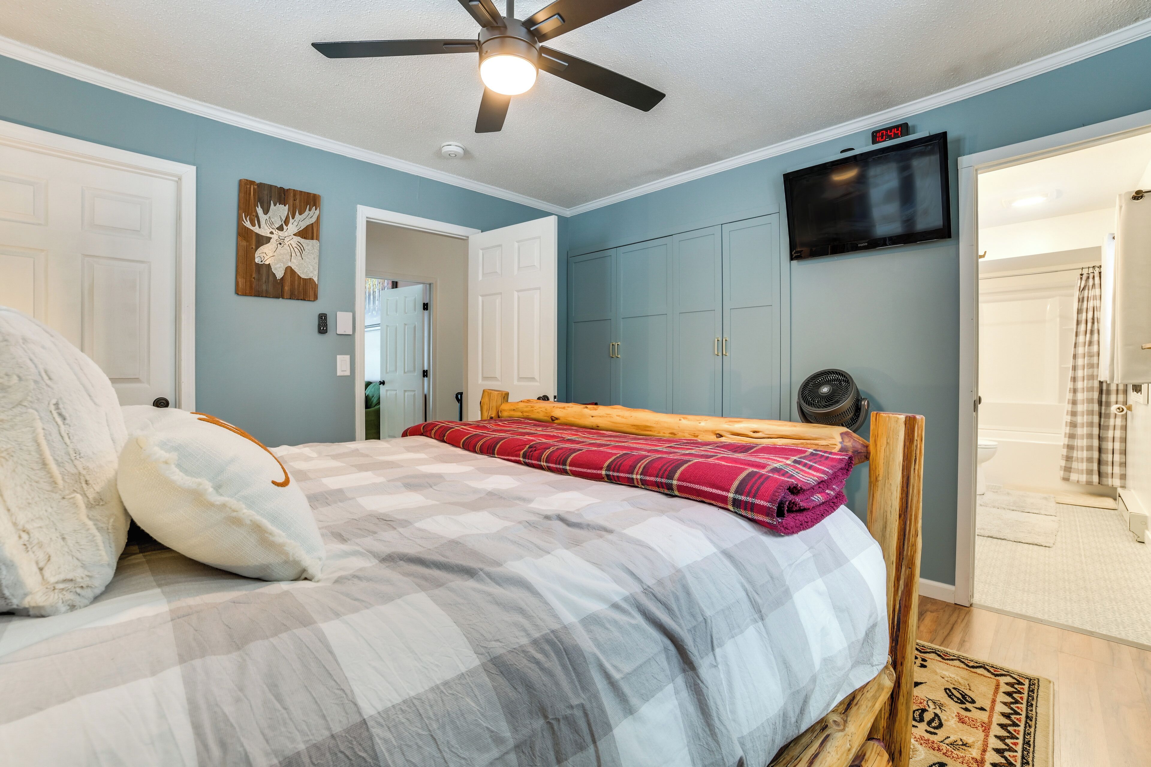 A bedroom with a bed, a ceiling fan, a mounted TV, and a door leading to a bathroom.