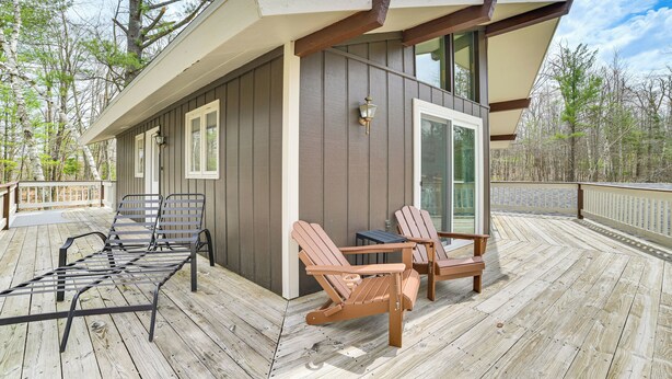 A wooden deck with Adirondack chairs and a small table, a covered area with a bench, and a building with large windows and a light fixture.