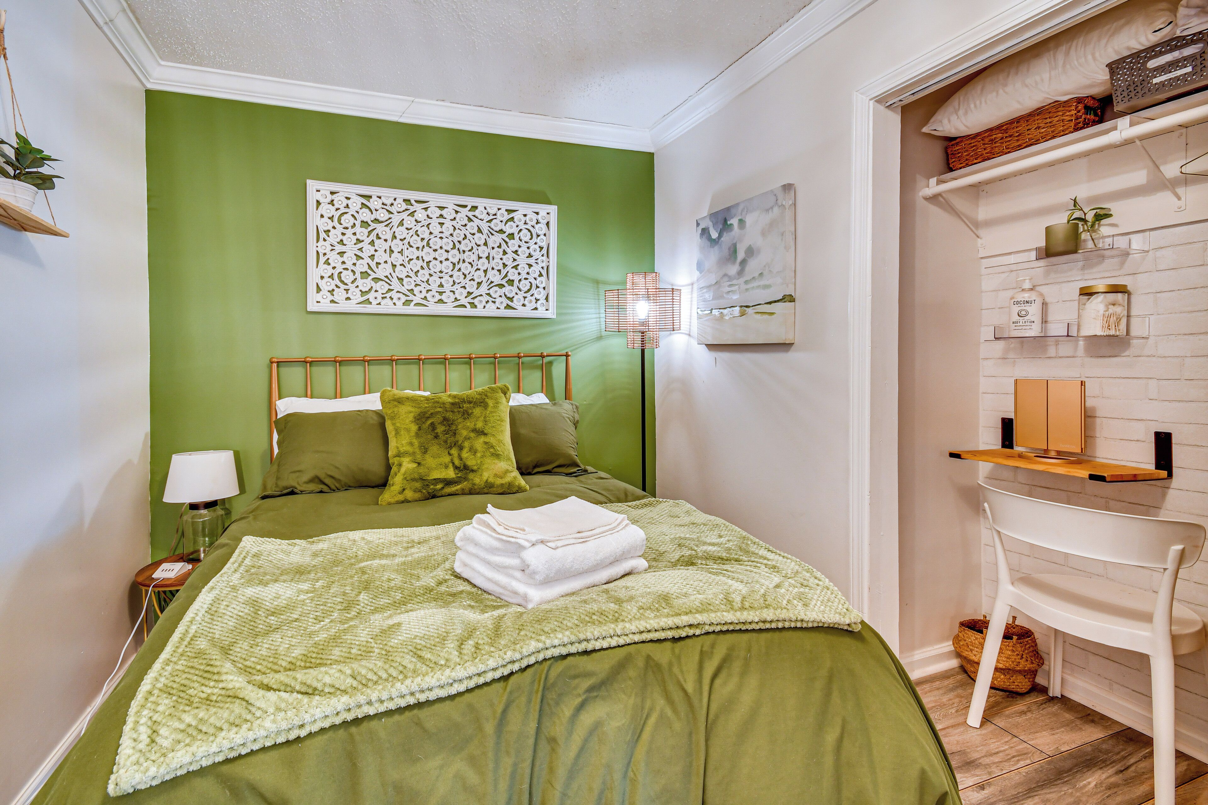A neatly made bed with a green bedspread, a white throw blanket, and a folded white towel. There is a small bedside table with a lamp and a power outlet.