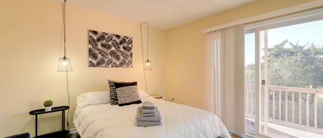 A bedroom with a large bed, two bedside tables, a ceiling fan, a window with a view of the outdoors, and a decorative wall piece.