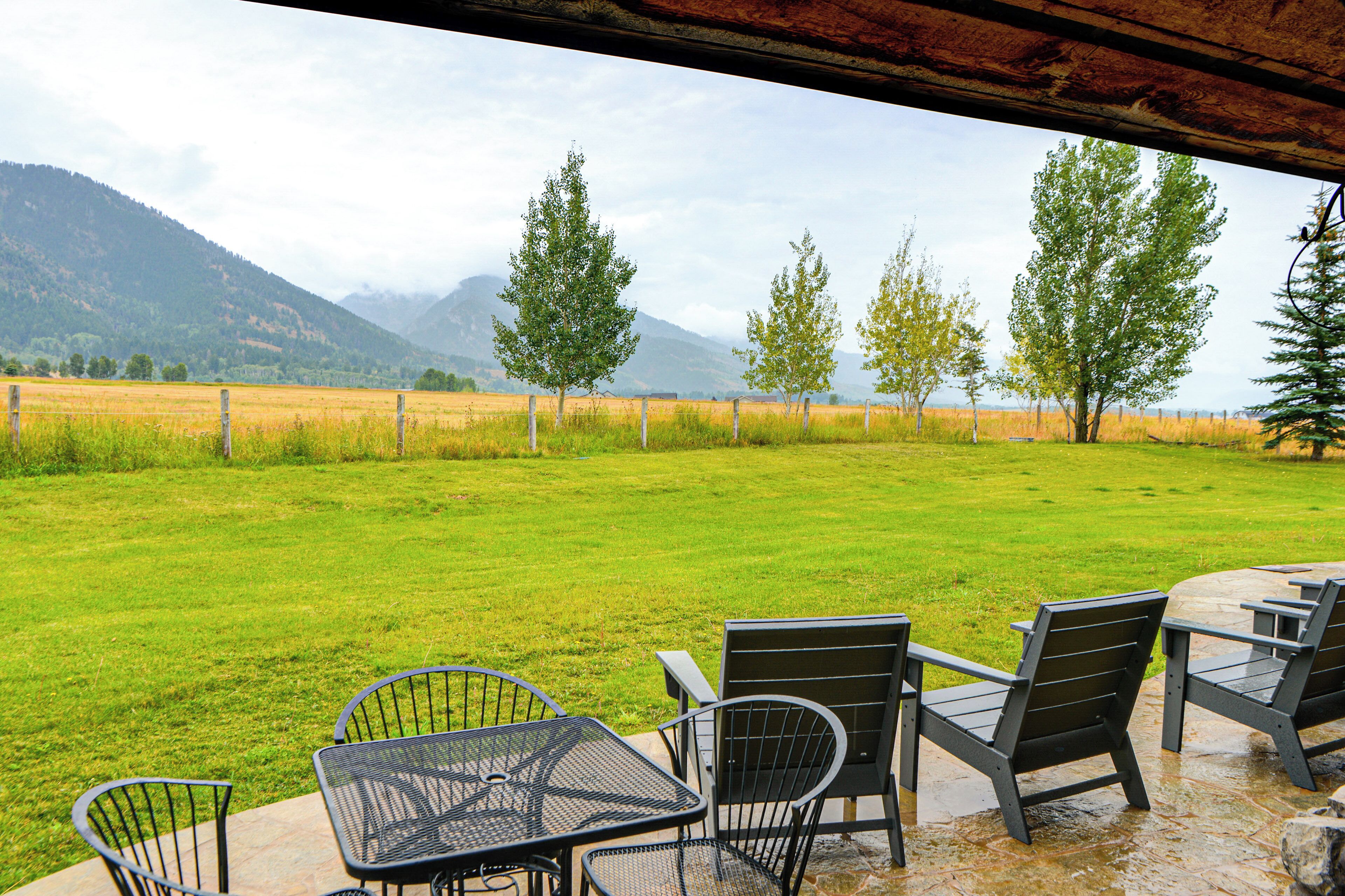An outdoor seating area with metal chairs and a table, overlooking a grassy field and mountains.