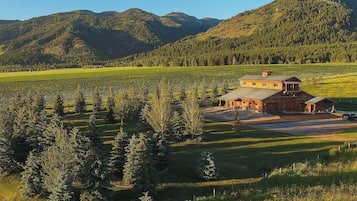 A rustic wooden barn with a large open area in front, surrounded by a forested landscape and mountains in the background.