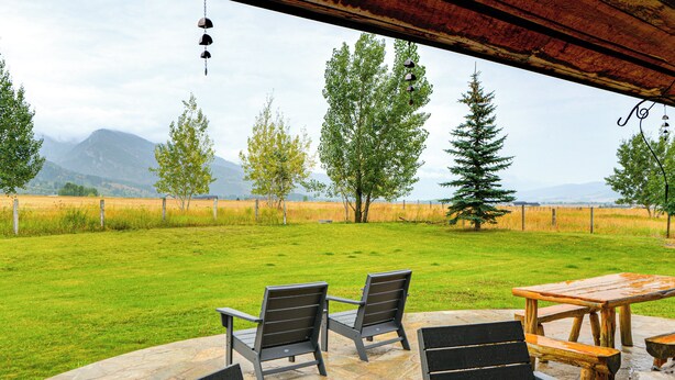 An outdoor patio with stone flooring, wooden furniture, and a view of a grassy field with trees and mountains in the distance.