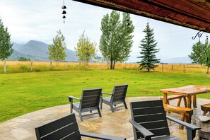Un patio al aire libre con suelo de piedra, mobiliario de madera y vistas a un campo cubierto de césped con árboles y montañas a lo lejos.