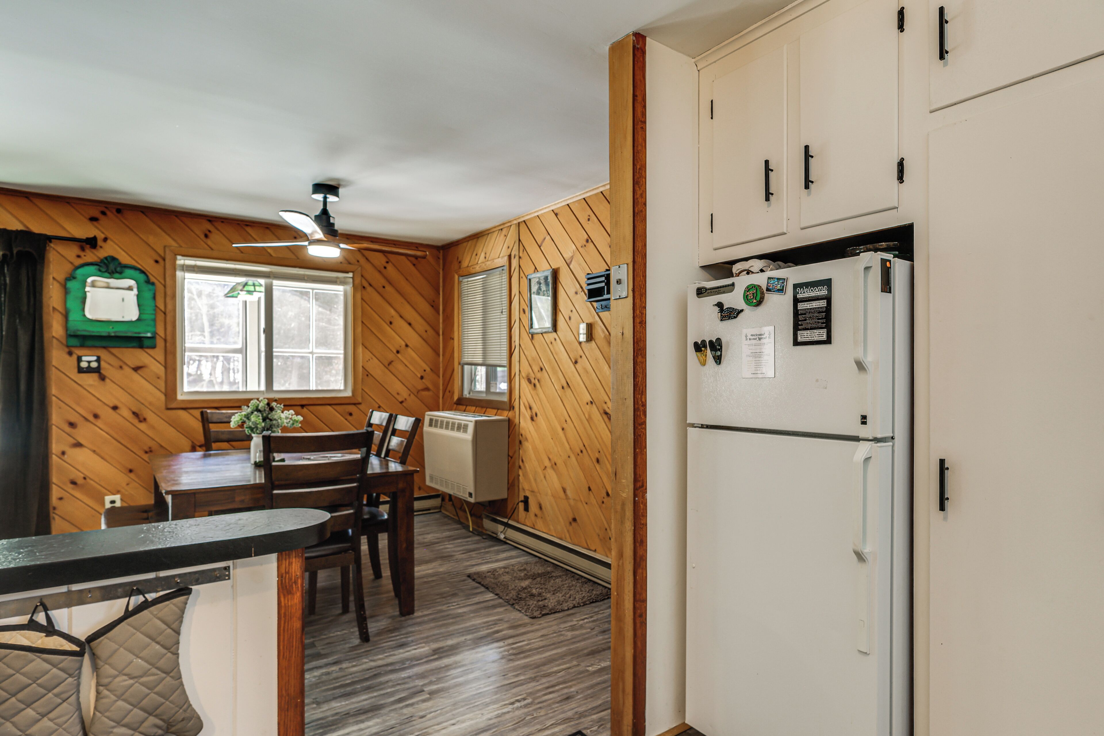 A kitchen with wooden walls, a refrigerator, a dining table with chairs, and a ceiling fan.