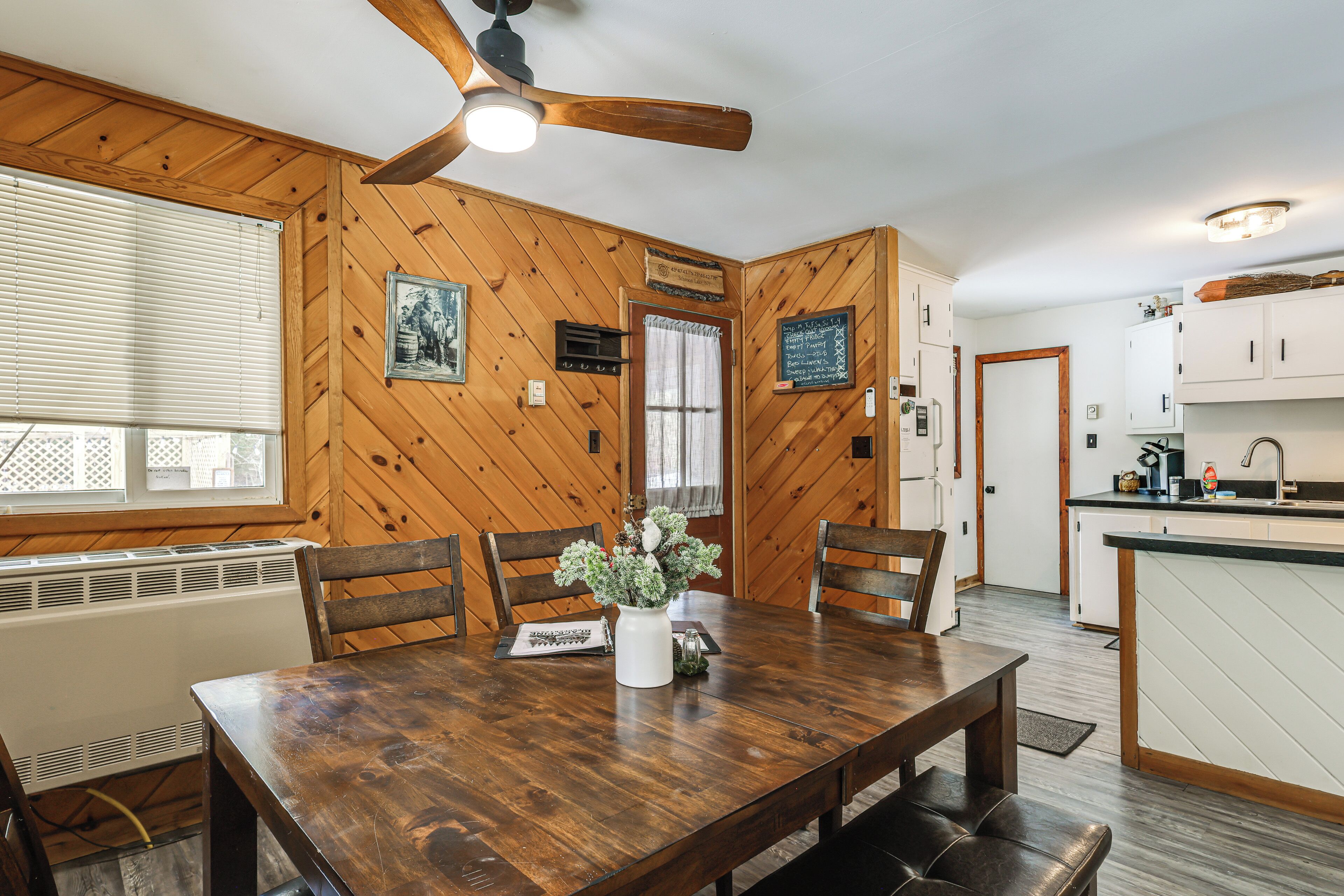 A wooden-paneled room with a dining table, chairs, a ceiling fan, a window with blinds, and a kitchen area with cabinets and a countertop.