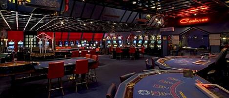 A casino with multiple blackjack tables, slot machines, and a neon sign for the cashier.