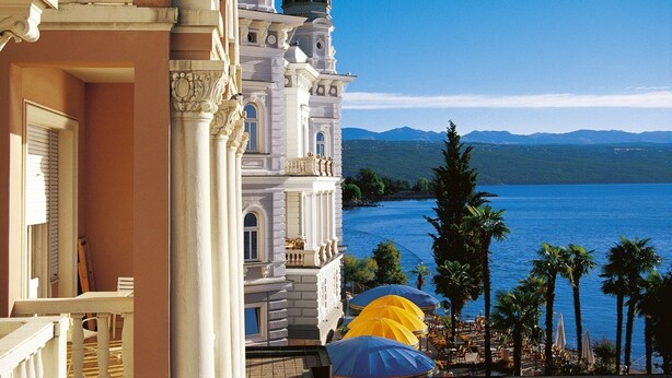 A grand building with classical architecture, overlooking a calm sea and distant mountains.