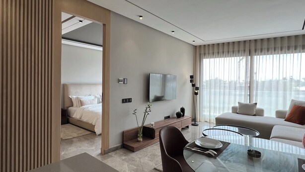 Una habitación de hotel moderna con cama, televisor montado en la pared, zona de comedor con mesa de cristal y un amplio ventanal con cortinas.